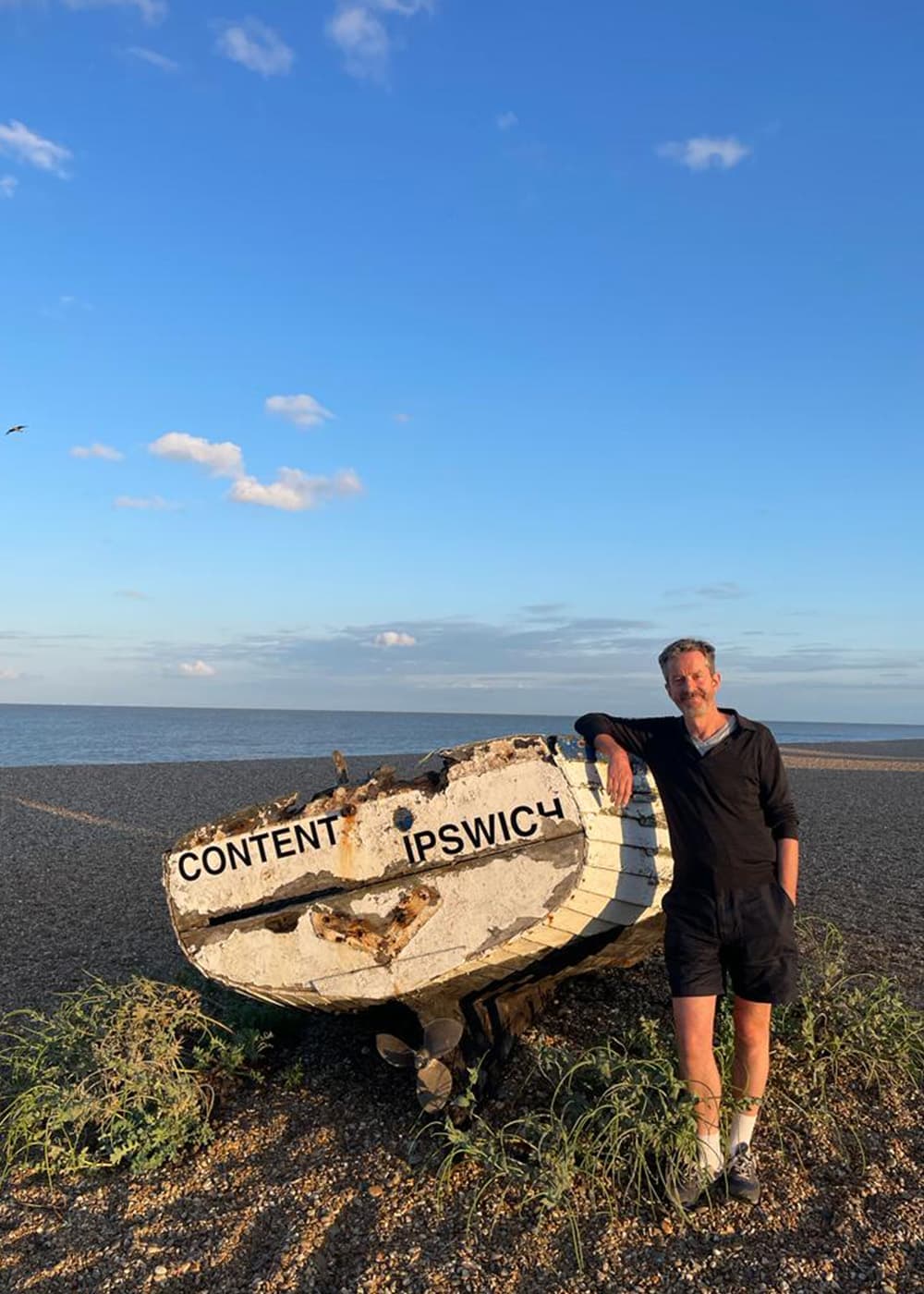 Alex Monroe by the coast in Ipswich leaning on an old boat.